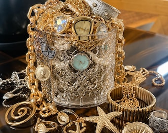 May include: A clear glass container filled with gold-toned jewelry including chains, watches, and charms. Surrounding the base are gold-toned rings, a starfish-shaped pendant, a shell-shaped piece with a pearl, and a textured metal bracelet.