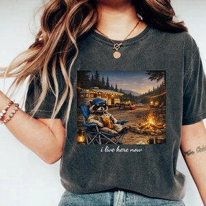 Puede incluir: Camiseta gris oscuro con un diseño de un mapache con gafas de sol y gorro azul, sentado en una silla de camping con una bebida. La imagen incluye una caravana, una fogata y el texto "i live here now".
