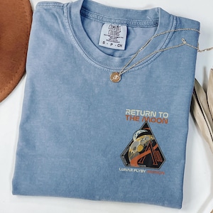 May include: A white ceramic mug with a black handle and the text "But First, Coffee" in a bold, black font. The mug is set against a blurred background, suggesting a cozy setting.