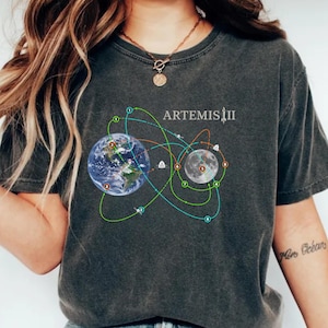 May include: Dark gray t-shirt featuring a graphic of Earth and the Moon connected by colorful lines, with the text "ARTEMIS II" above. The Earth and Moon are detailed with realistic imagery. The shirt has a crew neck and short sleeves.
