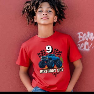 以下が含まれることがあります： 青いモンスター トラックのグラフィック、数字の9、および「BIRTHDAY BOY Taylor」のテキストが特徴の赤いTシャツ。 シャツは半袖で、赤い壁の前で人が着用しています。