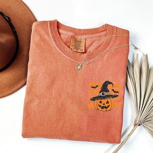 May include: A burnt orange short-sleeved t-shirt featuring a graphic of a jack-o'-lantern wearing a black witch hat, accented by small black bats and horizontal orange stripes. The shirt has a crew neckline and a Comfort Colors brand label.
