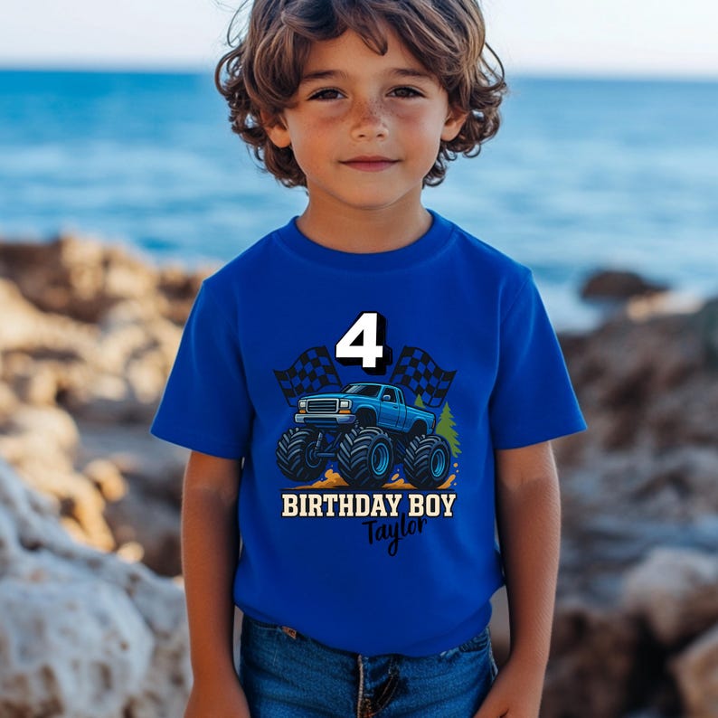 以下が含まれることがあります： モンスターカーのグラフィック、数字の4、そして「BIRTHDAY BOY Taylor」の文字が入った青いTシャツ。グラフィックには、チェッカーフラッグと大きなタイヤの青いトラックが含まれています。Tシャツは幼い子供が着ています。