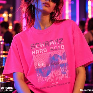 May include: A neon pink short-sleeve t-shirt featuring a graphic print with the text SCHRANZ HARD ACID and HARD TECHNO / RAVE UNIT. The design includes a pixelated blue and white landscape illustration and the text BPM 155 with an arrow.
