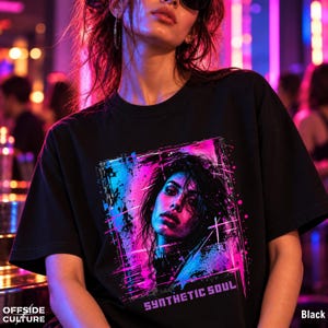 May include: A black t-shirt featuring a graphic print of a person's face in vibrant neon pink and blue colors. The design includes a rectangular frame with abstract splatter effects and the text "SYNTHETIC SOUL" printed in a bold font at the bottom.