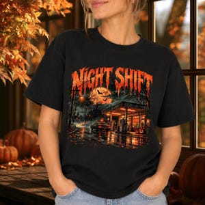 May include: A black short-sleeve t-shirt featuring a graphic print of a gas station at night under a full moon. The text NIGHT SHIFT appears in a dripping orange font above the scene, which includes a car, trees, and glowing pumpkins.