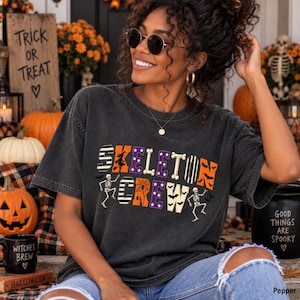 May include: A charcoal gray short-sleeve t-shirt featuring the text SKELETON CREW in large, multi-patterned block letters. The design includes orange, purple, and black stripes, spider webs, and bats, with two dancing skeleton illustrations.