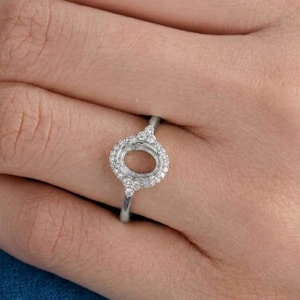 Puede incluir: Un anillo de tono plateado con un engaste central de forma ovalada rodeado por un halo de pequeñas piedras claras y brillantes. La banda es delgada y metálica, mostrando un diseño delicado que rodea el dedo con un acabado pulido.