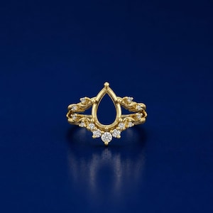May include: A gold ring guard featuring a central pear-shaped open space framed by a textured, branch-like metal band. The band is adorned with small, round, clear stones set along the lower curve and sides, creating a symmetrical, decorative design.