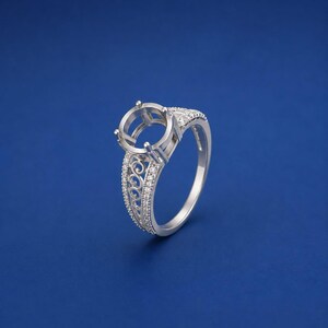 May include: A silver-toned ring setting featuring a circular prong mount for a centre stone. The band is decorated with intricate scrollwork patterns and small, clear pavé-set stones along the sides, creating a detailed and ornate appearance.