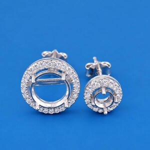 Peut inclure: Deux boucles d'oreilles à tige de couleur argentée avec un design circulaire en halo. Chaque boucle présente un sertissage central vide à griffes entouré d'un anneau de petites pierres rondes transparentes. Elles ont des fermoirs à tige.