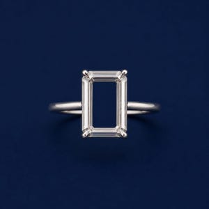 Könnte beinhalten: Ein silberfarbener Ring mit einem rechteckigen, offenen Rahmendesign. Die Mitte des Rings ist hohl und wird von vier geraden, metallischen Stäben eingerahmt, die an abgerundeten Ecken aufeinandertreffen und eine klare, geometrische Rechteckform bilden.