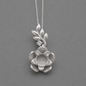 Puede incluir: Un collar de tono plateado con un colgante en forma de flor de cinco pétalos con un engaste circular central. Sobre la flor, un tallo curvado con tres hojas y una pequeña piedra redonda transparente está unido a una cadena delgada.