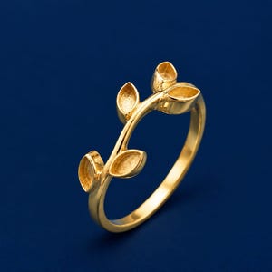 Puede incluir: Un anillo de tono dorado con una banda delgada que se ramifica en un diseño de enredadera. El anillo está adornado con cuatro engastes pequeños, huecos y con forma de hoja, cada uno diseñado para sostener una piedra o un adorno.