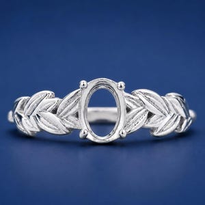 Puede incluir: Un anillo de plata con un engaste central ovalado de garras diseñado para sostener una piedra preciosa. La banda presenta un patrón en relieve detallado de hojas superpuestas que se extienden desde el engaste central a ambos lados.