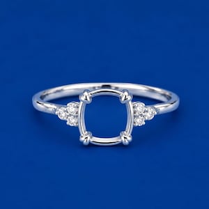 May include: A silver-toned ring setting featuring a central oval-shaped prong mount designed to hold a gemstone. The band is accented on both sides by a cluster of three small, clear, round-cut stones set into the metal shank.