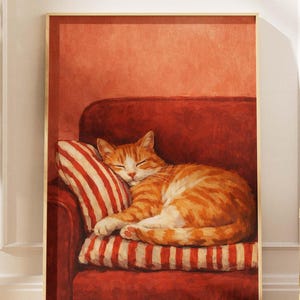 May include: A framed print of a ginger cat sleeping on a red sofa. The cat is curled up on a striped pillow. The background is a solid, warm red color.