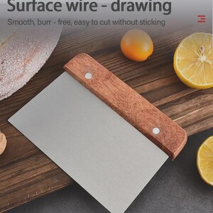 May include: A stainless steel dough scraper with a wooden handle. The text "Surface wire - drawing" and "Smooth, burr - free, easy to cut without sticking" are visible. The scraper is on a wooden surface with a lemon and orange.