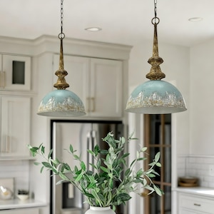 Pode incluir: Duas luminárias pendentes com acabamento azul e dourado envelhecido penduradas em correntes. As luminárias têm formato de cúpula e estão suspensas sobre uma cozinha branca. Um vaso de galhos verdes e frondosos está em primeiro plano.