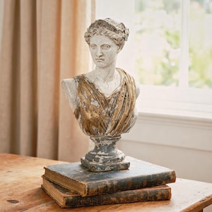 May include: A weathered bust of a woman with a gold-toned draped garment and a laurel wreath. The sculpture is mounted on a pedestal and sits atop two antique books. The bust is painted in shades of white, gray, and gold.