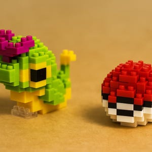 May include: A green and yellow block figure of a caterpillar-like creature with a pink headpiece, next to a red, white, and black block ball. The toys are on a tan surface.