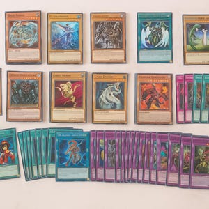 May include: A collection of trading cards featuring various fantasy creatures and characters. The cards have colorful illustrations and text, with a mix of green, purple, and tan card backs. The cards are arranged on a white surface.