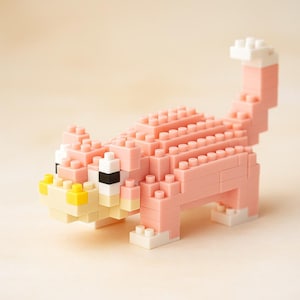 May include: A pink cat figurine constructed from interlocking building blocks. The cat features white, yellow, and black accents on its face and paws. The tail is raised, and the body is made of stacked blocks.