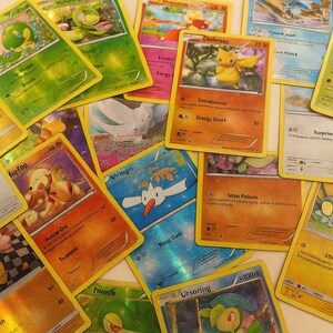 May include: A collection of colorful Pokémon trading cards, featuring various characters and designs. The cards are scattered across a light-colored surface, showcasing a variety of illustrations and text elements. The cards have yellow borders and are in good condition.