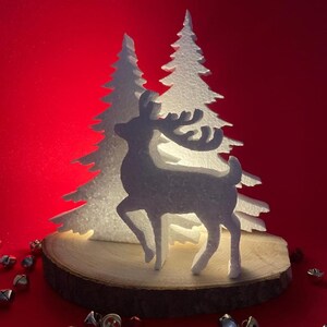 Può includere: Una silhouette bianca di renna su una base di legno con due alberi di Natale bianchi su sfondo rosso. Campane d'argento sparse intorno alla base. La scena è illuminata da una calda luce, creando un'atmosfera festosa.