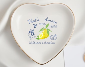 May include: A white heart-shaped ceramic trinket dish with a gold-toned rim. The centre features a graphic of yellow lemons, blue wedding rings, and a champagne bottle with the text That's Amore, 2026, Salut, and the names William & Amelia.