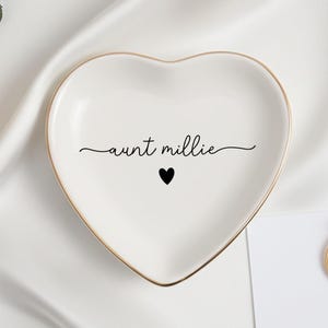 May include: A heart-shaped white ceramic ring dish with a gold rim, inscribed with "aunt millie" and a black heart. A gold ring and flowers are also visible.