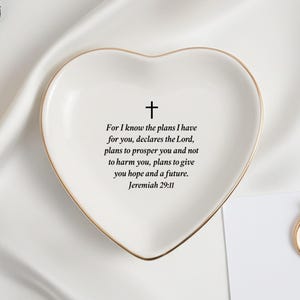 Peut inclure: Un vide-poches en céramique blanc en forme de cœur avec un bord doré, avec une croix et le verset biblique Jérémie 29:11. Une bague en or et une carte avec "MR. & MRS. MIL" sont également visibles.