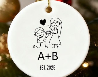 Puede incluir: Un adorno navideño de cerámica blanca con un dibujo lineal negro de una escena de propuesta de matrimonio. Un hombre se arrodilla ante una mujer, ofreciendo flores. El adorno tiene las iniciales "A+B" y el año "EST.2025" impresos en negro.