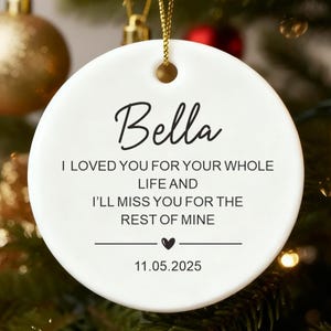 May include: A white ceramic Christmas ornament with the name "Bella" in cursive script. The ornament includes the text "I LOVED YOU FOR YOUR WHOLE LIFE AND I'LL MISS YOU FOR THE REST OF MINE" and the date "11.05.2025". The ornament hangs on a gold string.