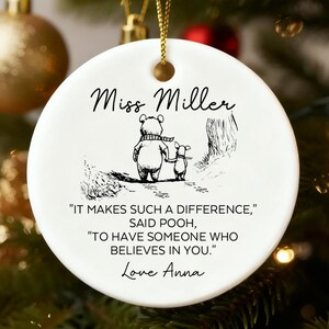 以下が含まれることがあります： 白いセラミック製のクリスマスオーナメント。くまのプーさんのイラストと「Miss Miller」の文字が描かれています。オーナメントにはプーさんの引用と「Love Anna」という名前が含まれています。