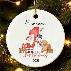 May include: A white ceramic Christmas bauble with a gold string, featuring a festive design. The bauble includes the text "Emma's 1st Christmas 2025" along with illustrations of Santa Claus, a snowman, and a gingerbread man. The bauble is hanging on a Christmas tree.