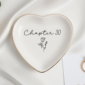 Op de afbeelding: Een hartvormige, witte keramische ringschaal met een gouden rand. De schaal heeft de woorden "Chapter 30" en een bloemmotief in zwart. Een gouden ring en een kaart met tekst zijn ook zichtbaar.