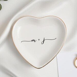 May include: A heart-shaped white ceramic ring dish with gold edging, inscribed with the initials "m + j" in cursive. A gold ring and a card with text are also present, set against a white satin background.