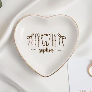 Op de afbeelding: Een hartvormige, witte keramische sieradenschaal met een gouden rand. De schaal heeft een afbeelding van tandheelkundige instrumenten en de naam "Sophia" in script. Een gouden ring en een kaart met tekst zijn ook zichtbaar.