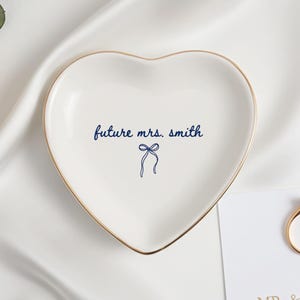 Peut inclure: Un vide-poche en céramique blanche en forme de cœur avec un bord doré. Le centre présente le texte bleu foncé "future mrs. smith" au-dessus d'un petit nœud bleu. Une bague dorée repose sur une carte blanche avec du texte partiel visible.
