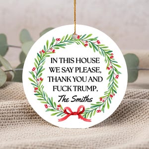 May include: A circular white Christmas decoration with a gold hanging string. It displays a green wreath with red berries and the text: "IN THIS HOUSE WE SAY PLEASE, THANK YOU AND FUCK TRUMP, The Smiths."