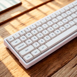 Może przedstawiać: Biała klawiatura mechaniczna z białymi klawiszami i czarnymi literami. Klawiatura znajduje się na drewnianej powierzchni. Klawisze zawierają litery, cyfry i znaki specjalne. Klawiatura ma kompaktową konstrukcję.