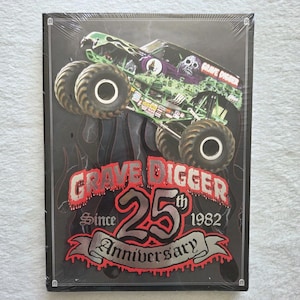 May include: A black and white Grave Digger 25th Anniversary collectible. The image features a monster truck with a green and black design, a skull, and the words "Grave Digger" and "Since 1982".