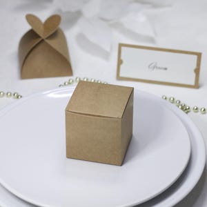 Peut inclure: Une petite boîte cadeau carrée en papier brun est posée sur une assiette blanche. Une autre boîte en forme de cœur et un carton de place avec le mot "Groom" sont également visibles. La scène suggère un mariage ou un événement.