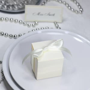 Peut inclure: Une petite boîte cadeau crème, nouée d'un ruban de satin, posée sur une assiette blanche. Une carte de place avec le nom "Miss Smith" est visible en arrière-plan, ainsi qu'un collier de perles argentées. La scène suggère un mariage ou un événement spécial.