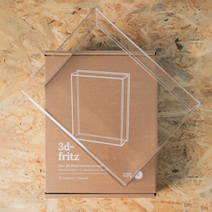 Op de afbeelding: Een doorzichtige acryl 3D-displaybox-inzetstuk geplaatst over een bruine kartonnen doos. De verpakking bevat een witte lijntekening van de rechthoekige vitrine, de tekst 3d-fritz en details die aangeven dat deze is voor objecten van 24x30 cm.