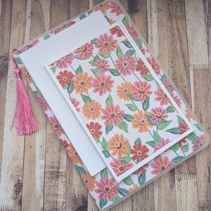 May include: A stationery set with a floral pattern of pink, orange, and red flowers on white. The set includes a notebook, a card, and a pink tassel. The notebook and card have white borders. The set is designed for writing.