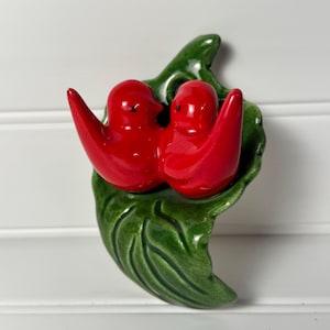 May include: A glossy ceramic wall plaque featuring two bright red birds perched together on a large, textured green leaf. The birds have smooth, rounded bodies and small black eyes, while the leaf displays detailed veins and a shiny finish.