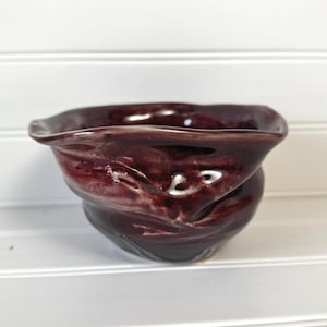 May include: A small, deep bowl with a glossy, dark burgundy glaze. The ceramic vessel features an irregular, wavy, and organic rim shape with a rippled texture along the exterior walls, creating a fluid and dynamic appearance in the light.