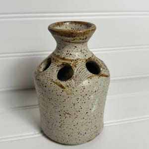May include: A speckled beige ceramic flower frog vase with a wide neck and three circular openings on the shoulder. The stoneware features a textured surface with dark brown flecks and a darker brown glazed rim, showcasing a hand-thrown appearance.
